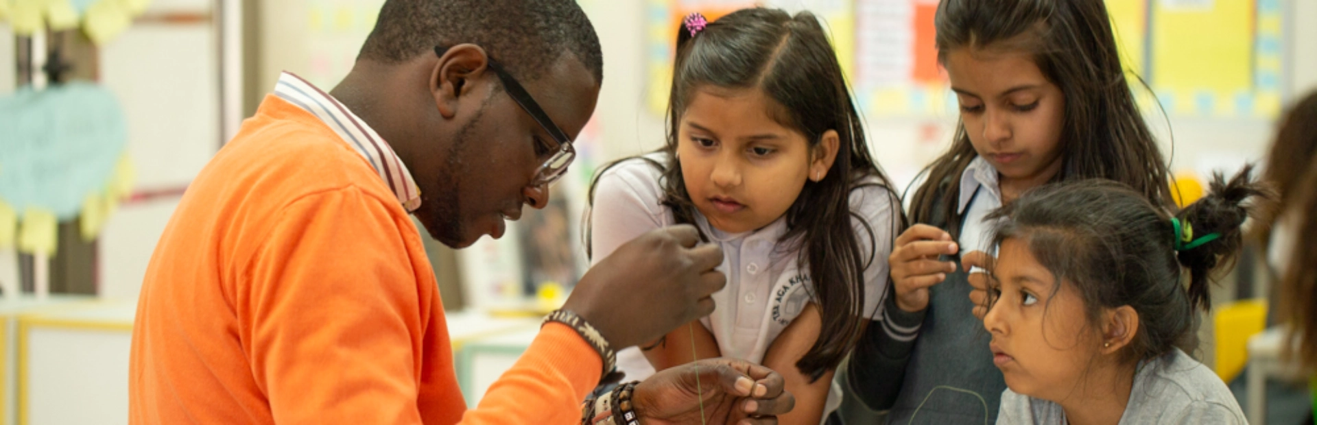  A teacher leans over a classroom table to assist a PYP student with a painting activity. The student attentively observes as the teacher demonstrates a technique with a paintbrush.