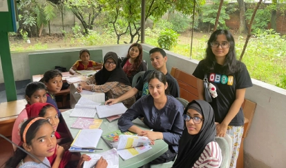 AKA Dhaka students and young children sit around tables in an outdoor learning area, engaged in an educational activity.