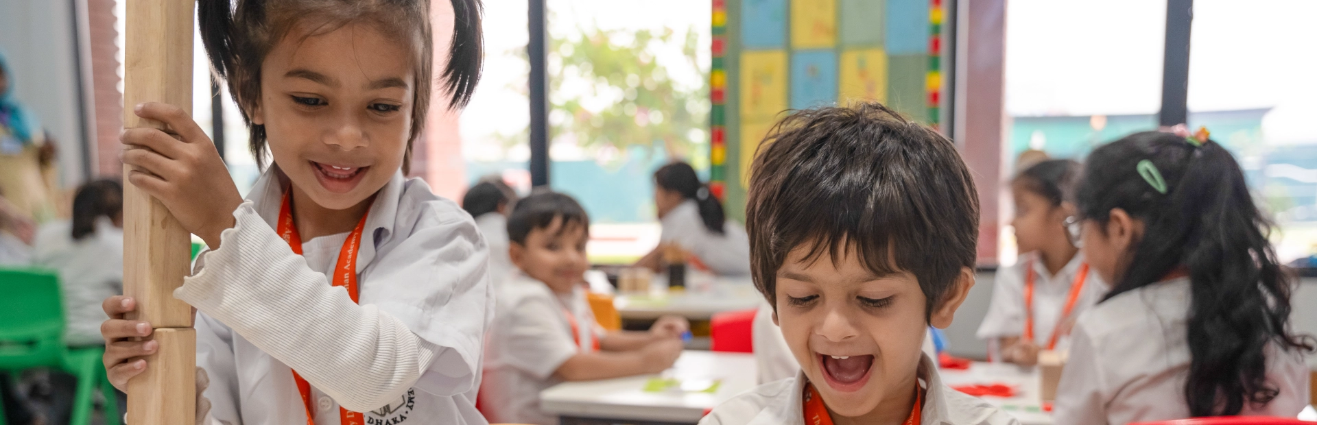  Young students at the Aga Khan Academy Dhaka smile and engage in classroom activities, wearing white uniforms and orange lanyards.