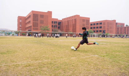 A student kicks a football on a grassy field near school buildings.