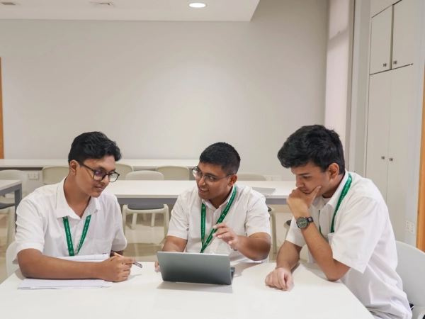 Three DP students sit together discussing their studies while using a laptop and notebooks at a classroom table.