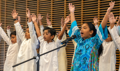Junior School students stand on stage, raising their hands while singing into a microphone. One student in a bright blue patterned outfit leads the song, surrounded by classmates dressed in white traditional attire.