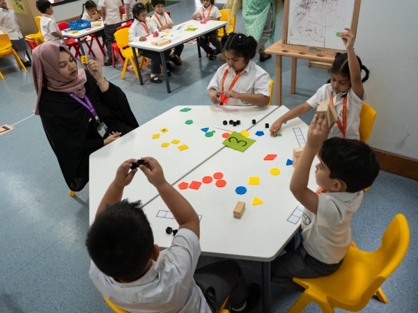A teacher and a group of young students sit around a classroom table using colourful blocks and shapes to learn numbers and patterns during an interactive maths lesson.