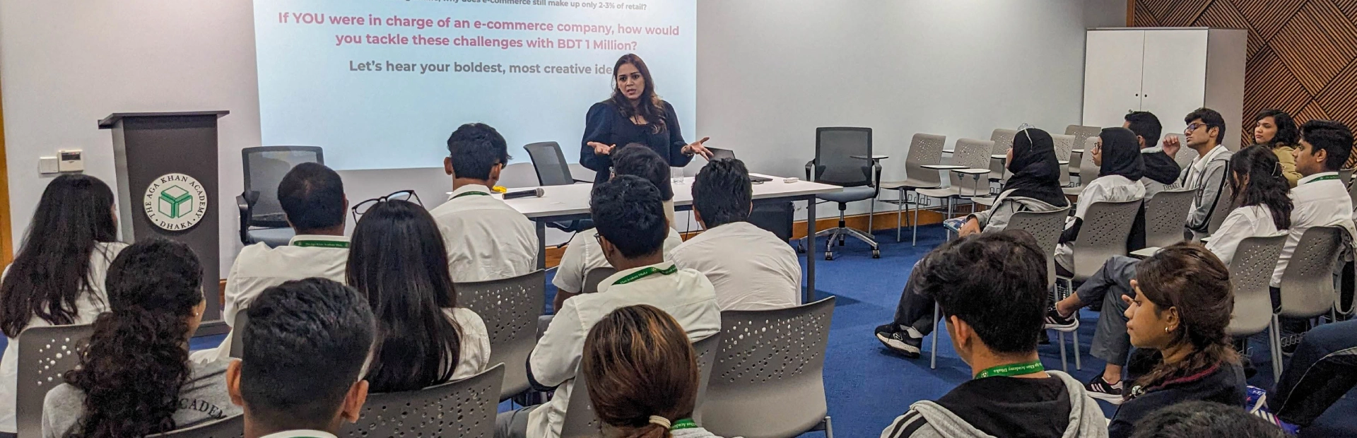 A group of students sitting in chairs with their backs to the camera. A woman is standing at the front of the room in front of a projection on the wall, addressing the students.
