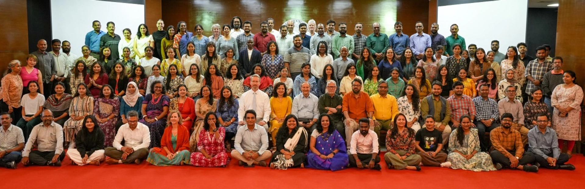 A woman sits at a desk smiling at the camera while a colleague works at a standing desk with multiple computer screens, in an office filled with colourful sticky notes and charts.