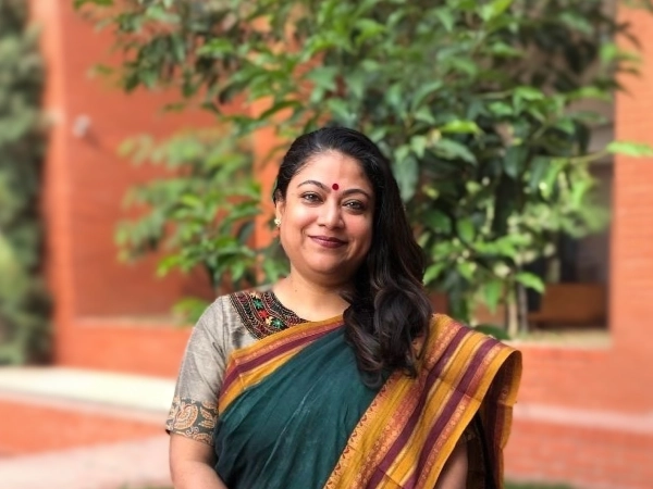 A teacher wearing a green and mustard saree stands smiling outdoors on the Aga Khan Academy Dhaka campus; she is the Lead Integrated Social Science Teacher.
