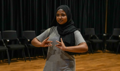 A student performs a dance pose during a drama class on stage, with black curtains and empty chairs in the background