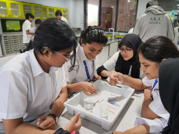 Four students in white uniforms work together around a tray containing test materials and beakers of water. They handle strips of paper and discuss observations, displaying curiosity and teamwork in a laboratory setting.