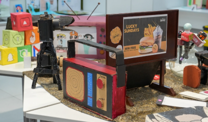 A display table features colourful student-made models, including a cardboard radio, camera, and advertisement board, surrounded by posters and craft cubes decorated with social media icons